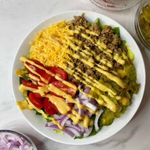Burger bowl with cooked grouond beef, shredded cheddar cheese, chopped pickles, sliced red onions, sliced romaine lettuce and halved grape tomatoes all drizzled with Burger Sauce Dressing.