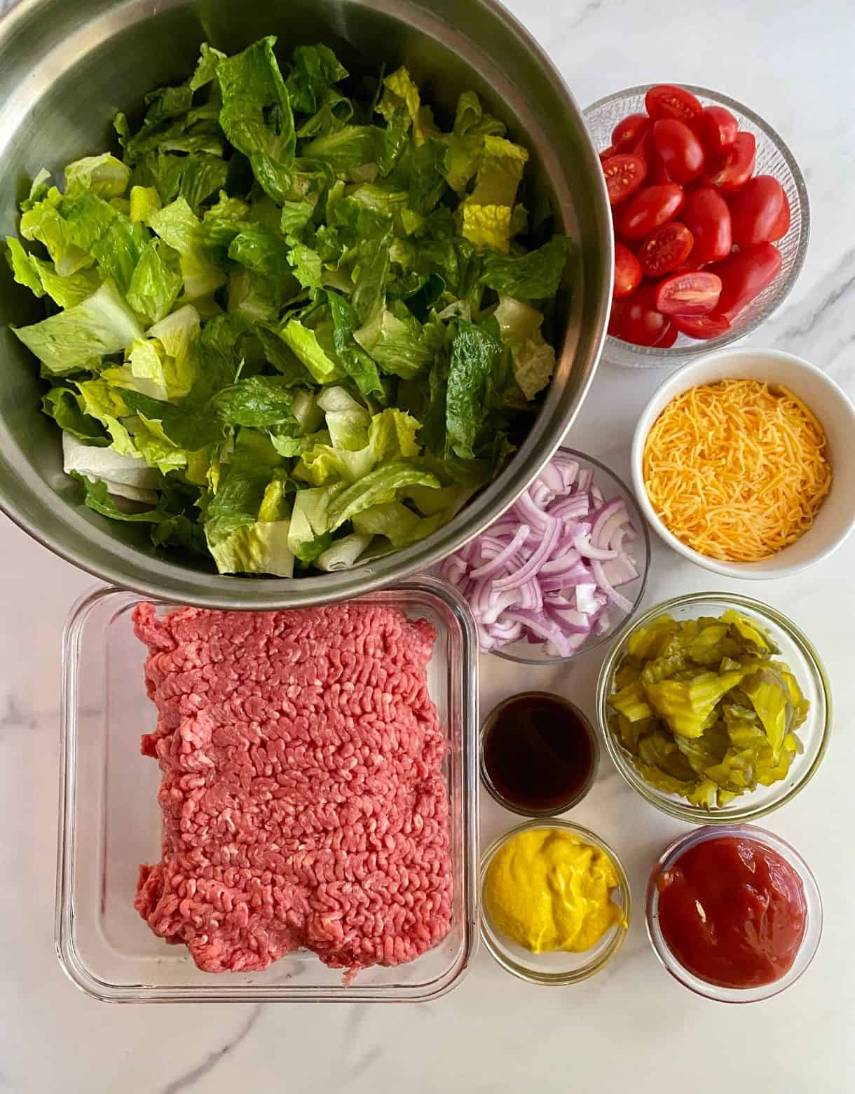 Ingredients for Burger Bowls. Chopped lettuce, raw ground beef, sliced red onion, halved grape tomatoes, shredded cheddar cheese, Worcestershire sauce, chopped pickled, mustard and ketchup.