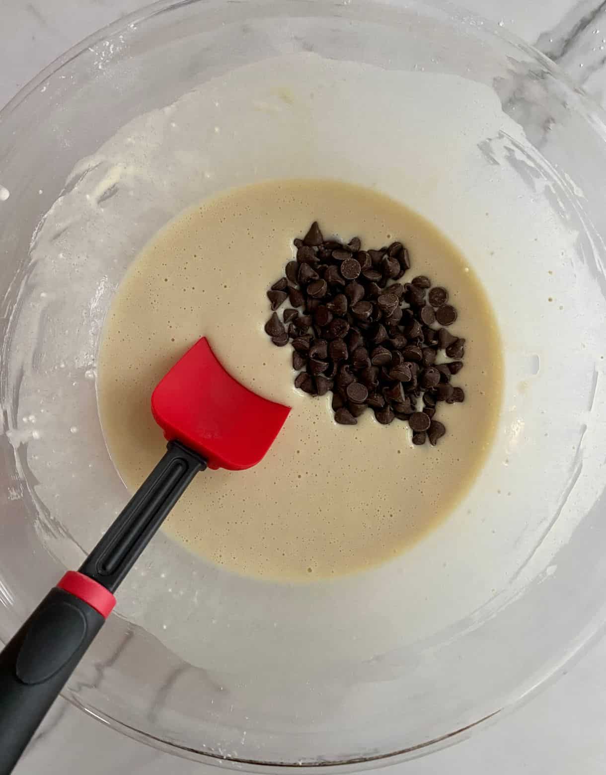 A bowl wih batter for Chocolate Chip Pancakes with a spoon and a pile of chocolate chips on top.