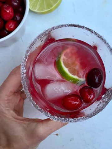 A hand holding a glass of Cranberry Margarita with a salted rim.