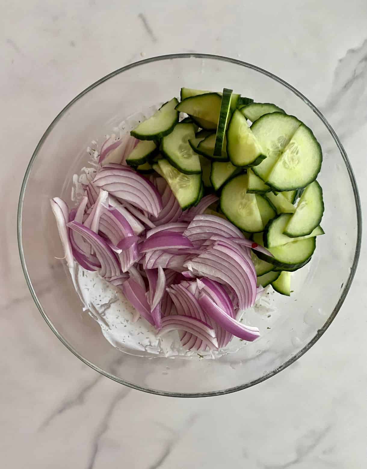 A bowl with dressing, sliced onions and diced cucumbers not mixed together.