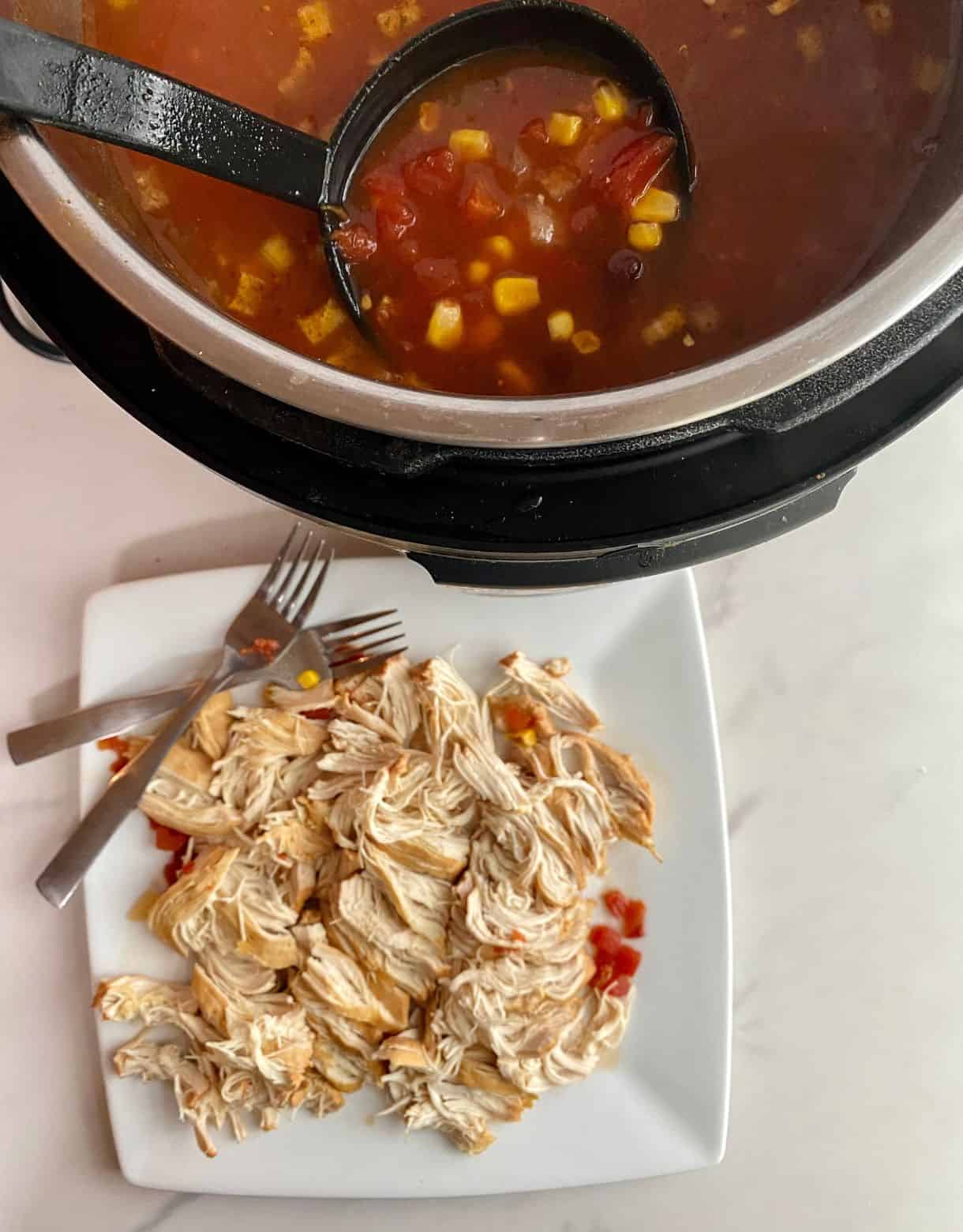 A slow cooker of cooked soup and a plate with shredded chicken breasts to be added to the soup.