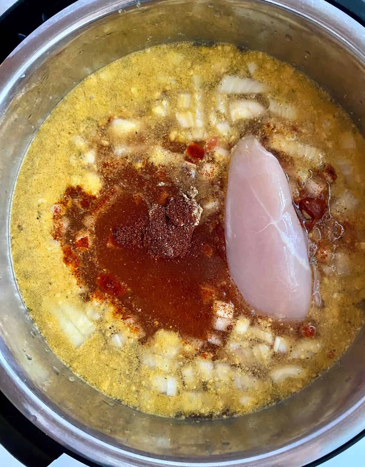 A slow cooker with chicken broth, diced onion, canned tomatoes, raw chicken breasts and spices added but not yet stirred.