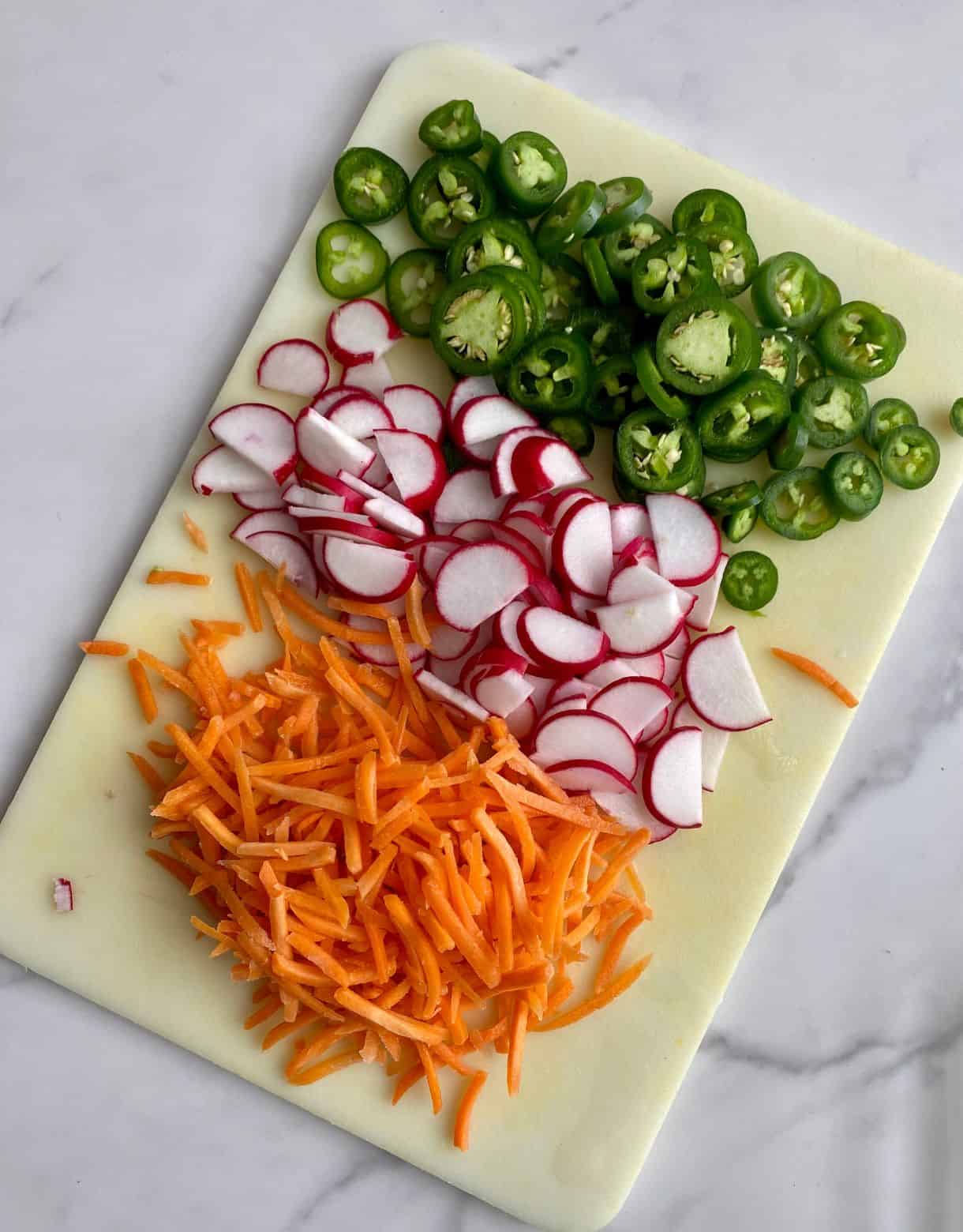 A cutting board with shredded carrots, sliced radishes and sliced jalapenos all on a cutting board.