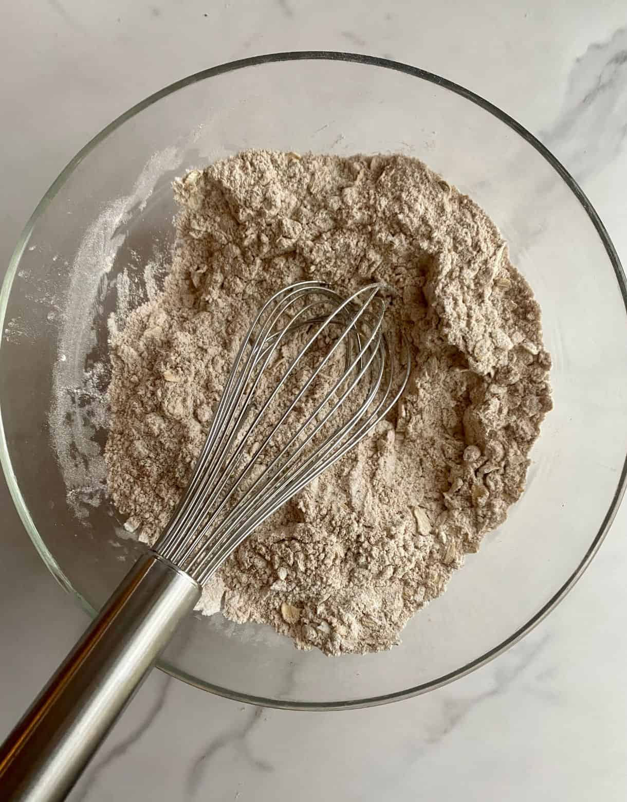 A bowl with flour, whole wheat flour and rolled oats and a wire whisk.