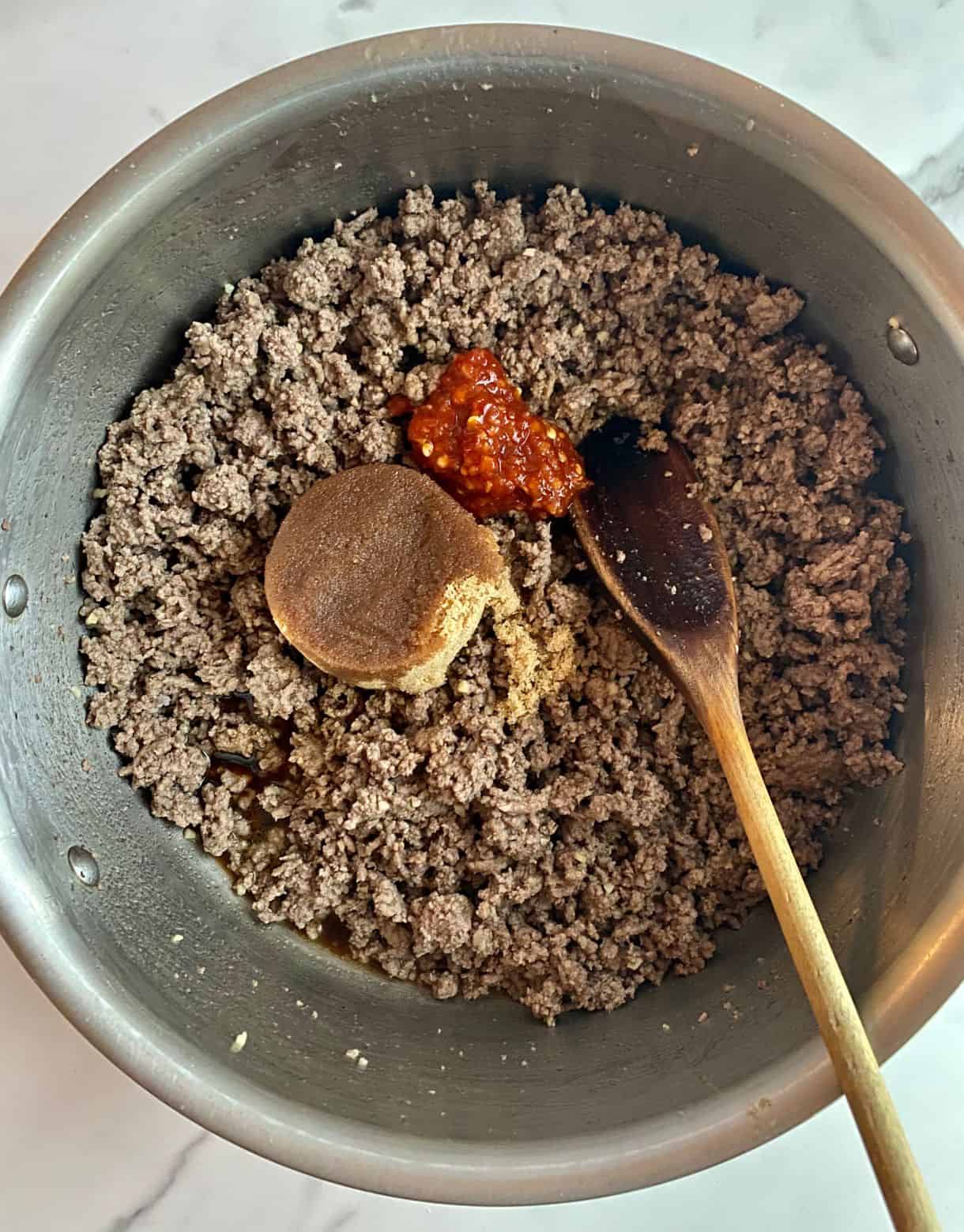 A skillet with cooked ground beef with brown sugar, soy sauce and chili garlic sauce added but not stirred in.