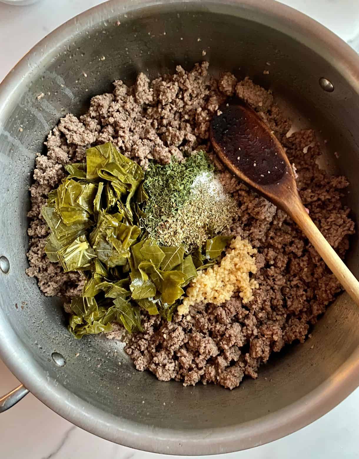 A pot with cooked and crumbled ground beef with chopped grape leaves, minced garlic and spices added but not yet stirred.