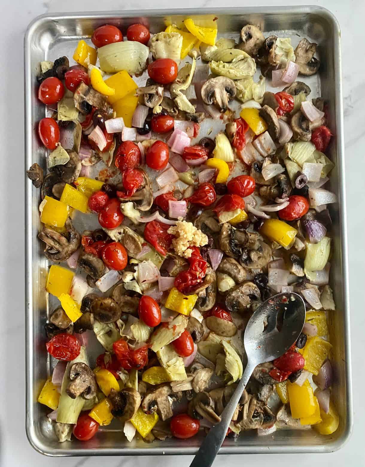 A sheet pan with half-roasted vegetables with minced garlic added but not yet stirred in.