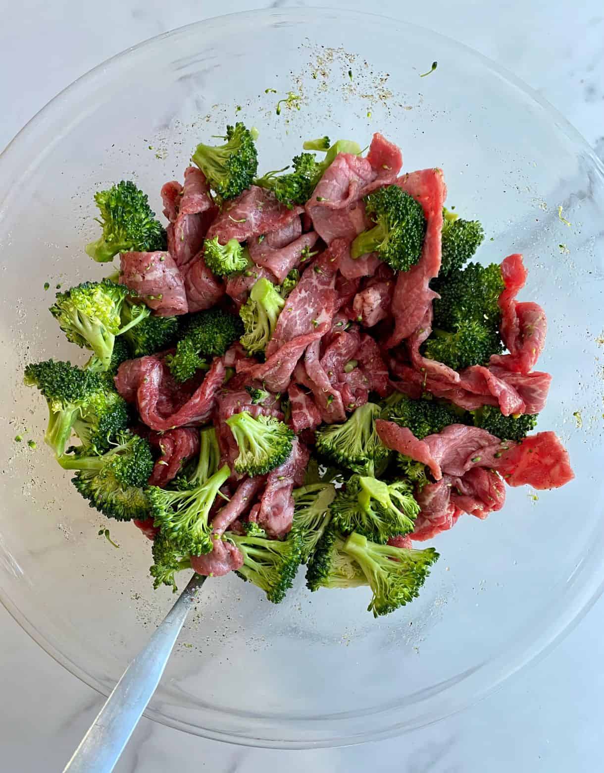A bowl with raw beef and broccoli mixed with olive oil, salt and pepper.