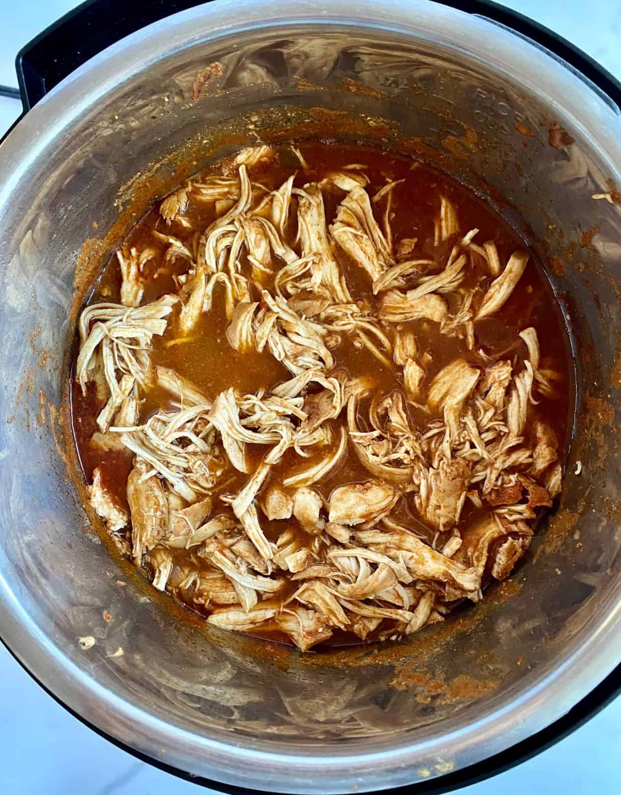 A slow cooker with cooked shredded chicken in a red salsa-based sauce.