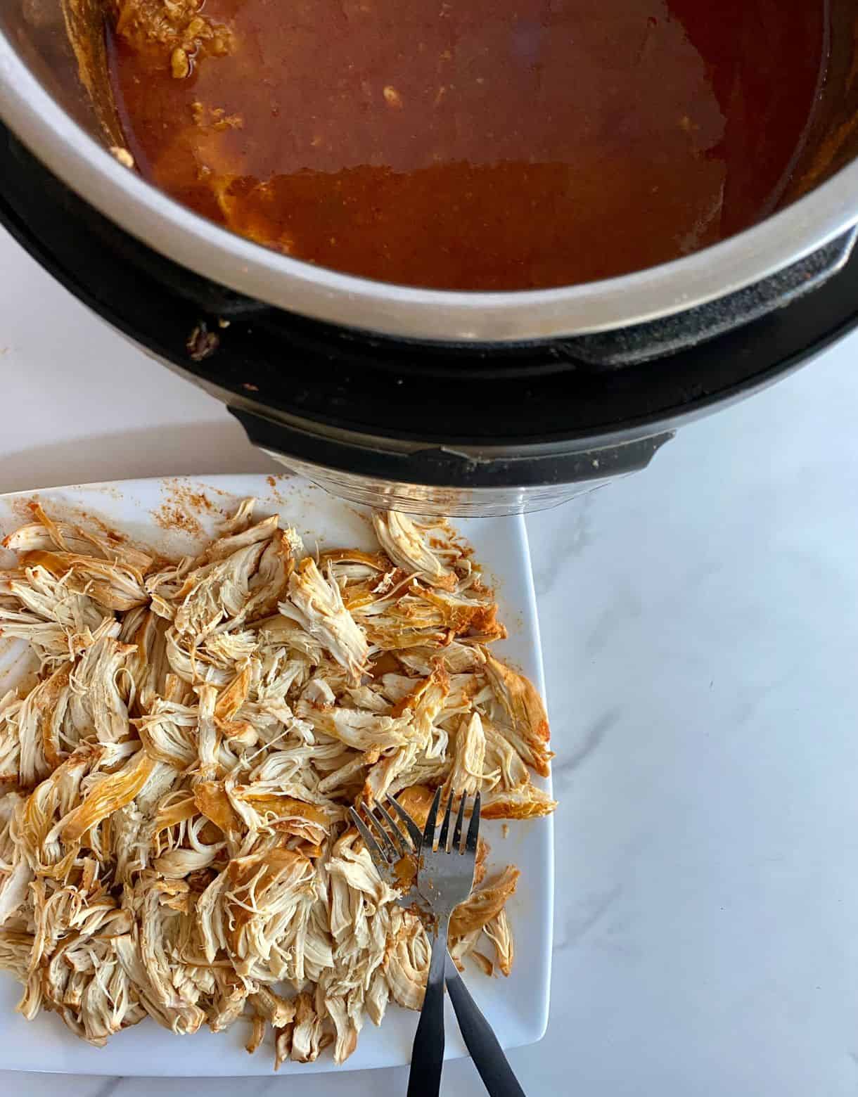 A plate with shredded chicken and two forks and the edge of the slow cooker with the red cooking juices.