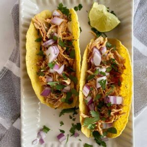 Two Slow Cooker Chicken Tacos on a plate with chopped raw and onion cilantro and a wedge of lime.