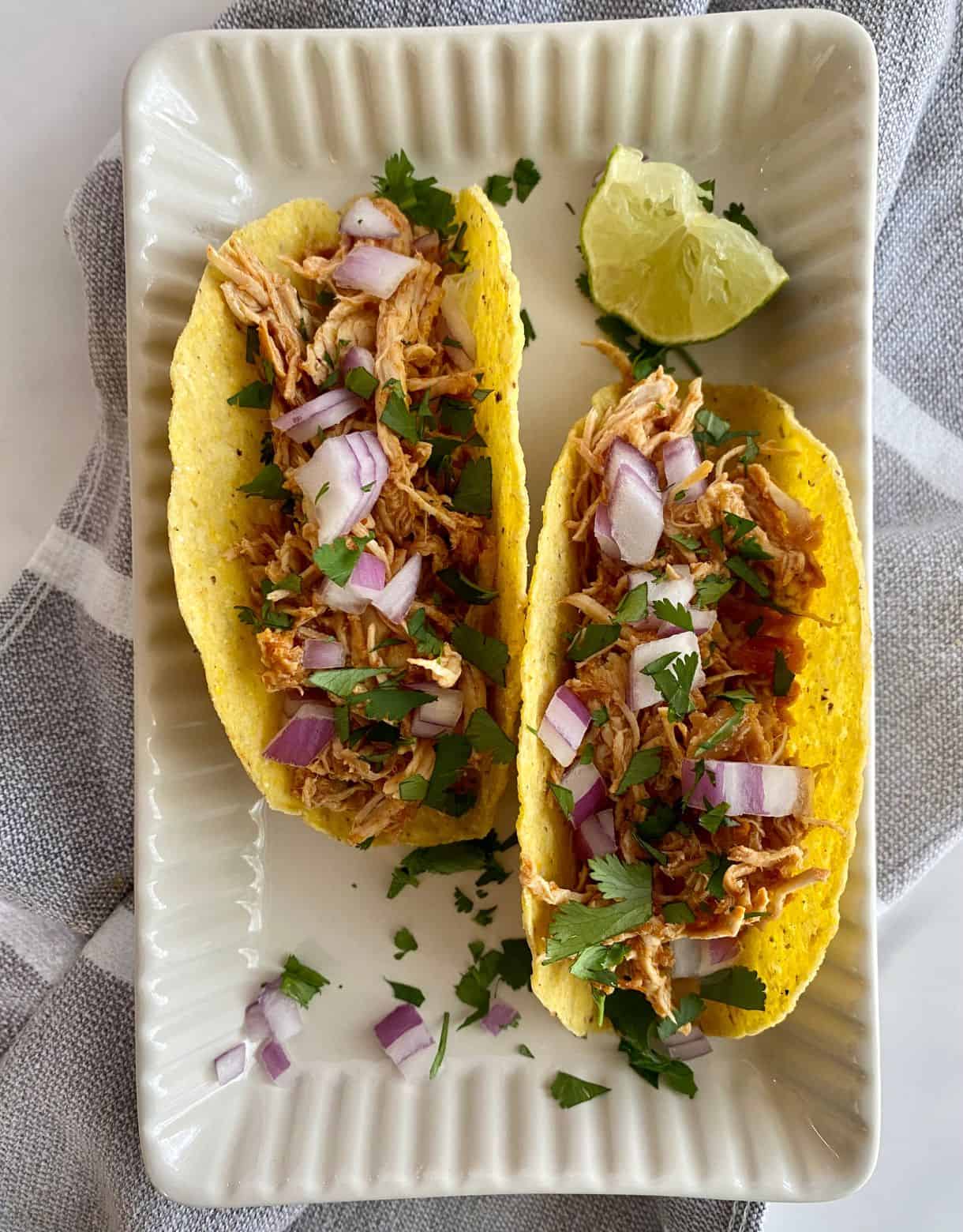 Two Slow Cooker Chicken Tacos on a plate with chopped raw and onion cilantro and a wedge of lime.