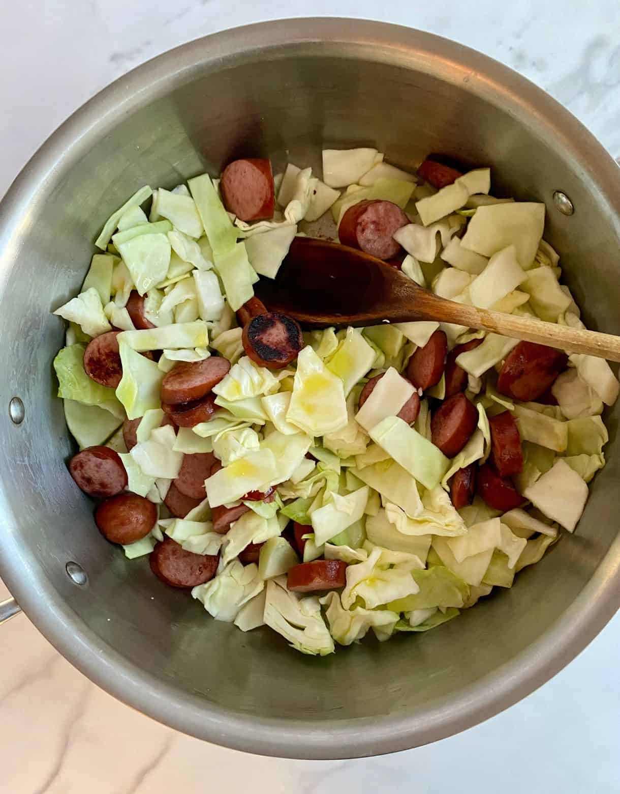 A skillet with browned sliced smoked sausage and chopped raw green cabbage with olive oil drizzled over top.
