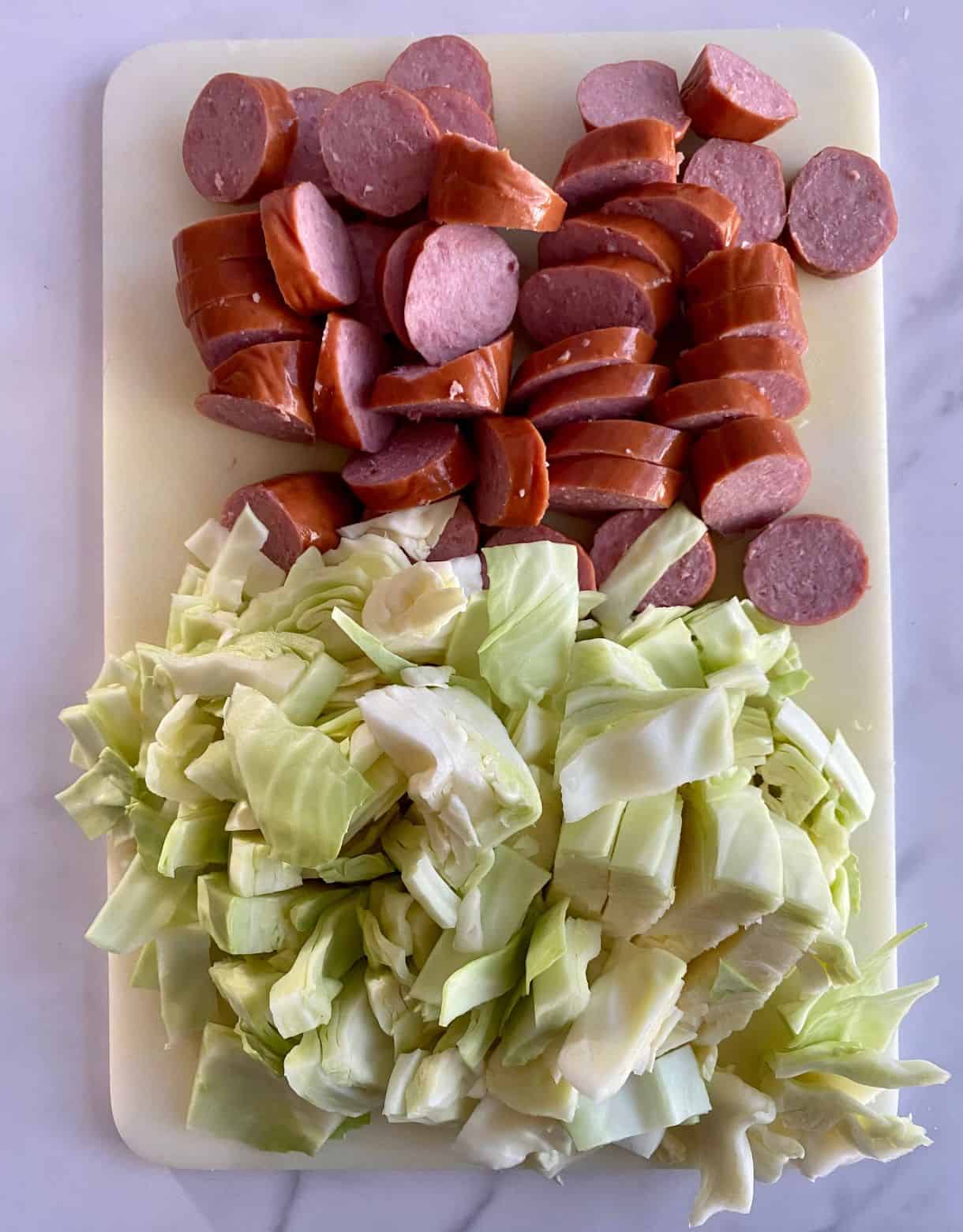 A cutting board with sliced smoked sausage and chopped cabbage.