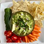 A plate with a bowl of Spicy Guacamole as well as sliced cucumbers, grape tomatoes, baby carrots and tortilla chips to dip in it. Plus the Healthy Mom Healthy Family logo.