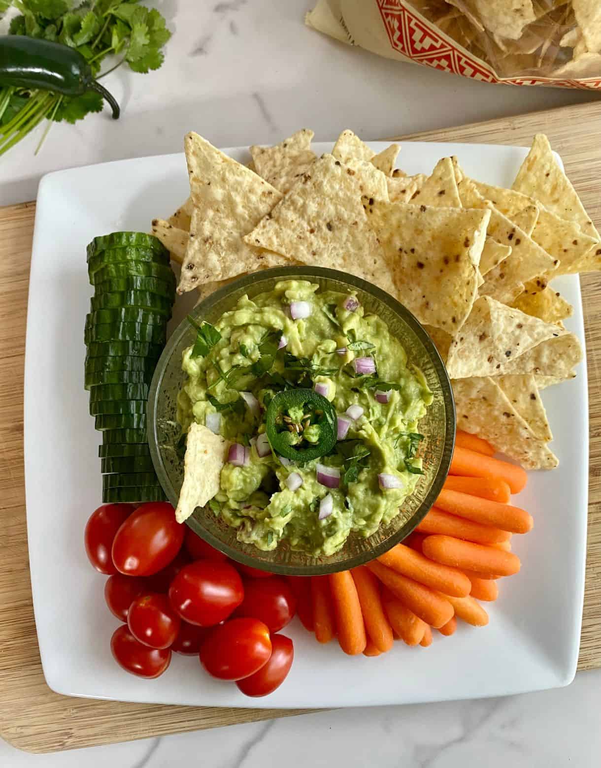 A plate with a bowl of Spicy Guacamole as well as sliced cucumbers, grape tomatoes, baby carrots and tortilla chips to dip in it.