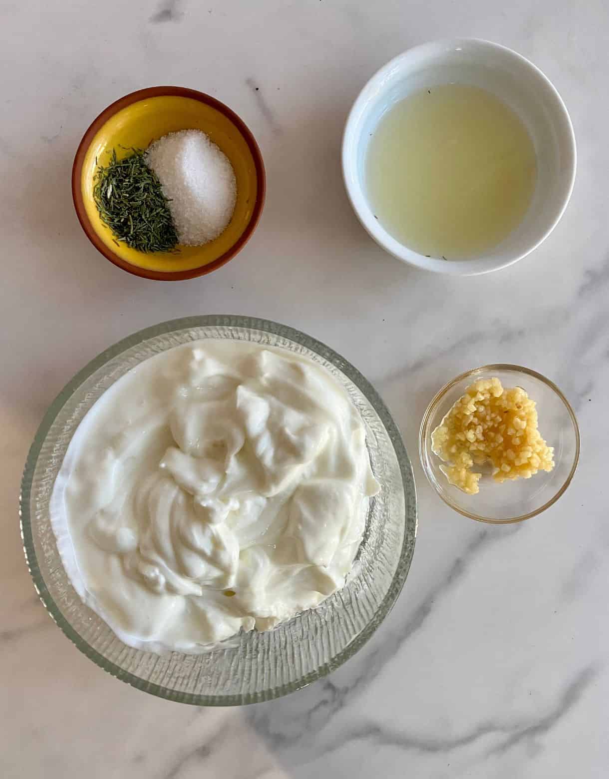 Bowls of plain Greek yogurt, minced garlic, lemon juice, salt and dried dill.