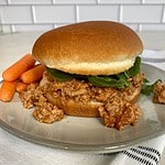A bun with Buffalo Sloppy Joe topped with arugula on a plate with carrots.