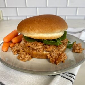 A bun with Buffalo Sloppy Joe topped with arugula on a plate with carrots.