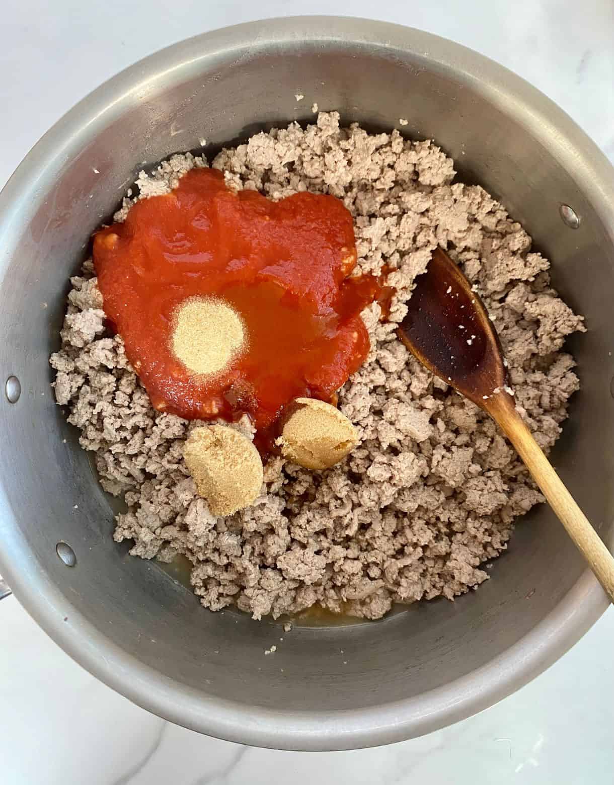 A pot with cooked and crumbled ground turkey with tomato sauce, brown sugar, garlic powder, vinegar, Worcestershire sauce and hot sauce added but not yet stirred.