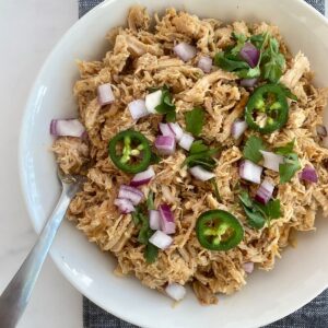 A bowl of cooked pork loin carnitas topped with sliced jalapenos and chopped onion and cilantro.