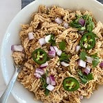 A bowl of cooked pork loin carnitas topped with sliced jalapenos and chopped onion and cilantro.