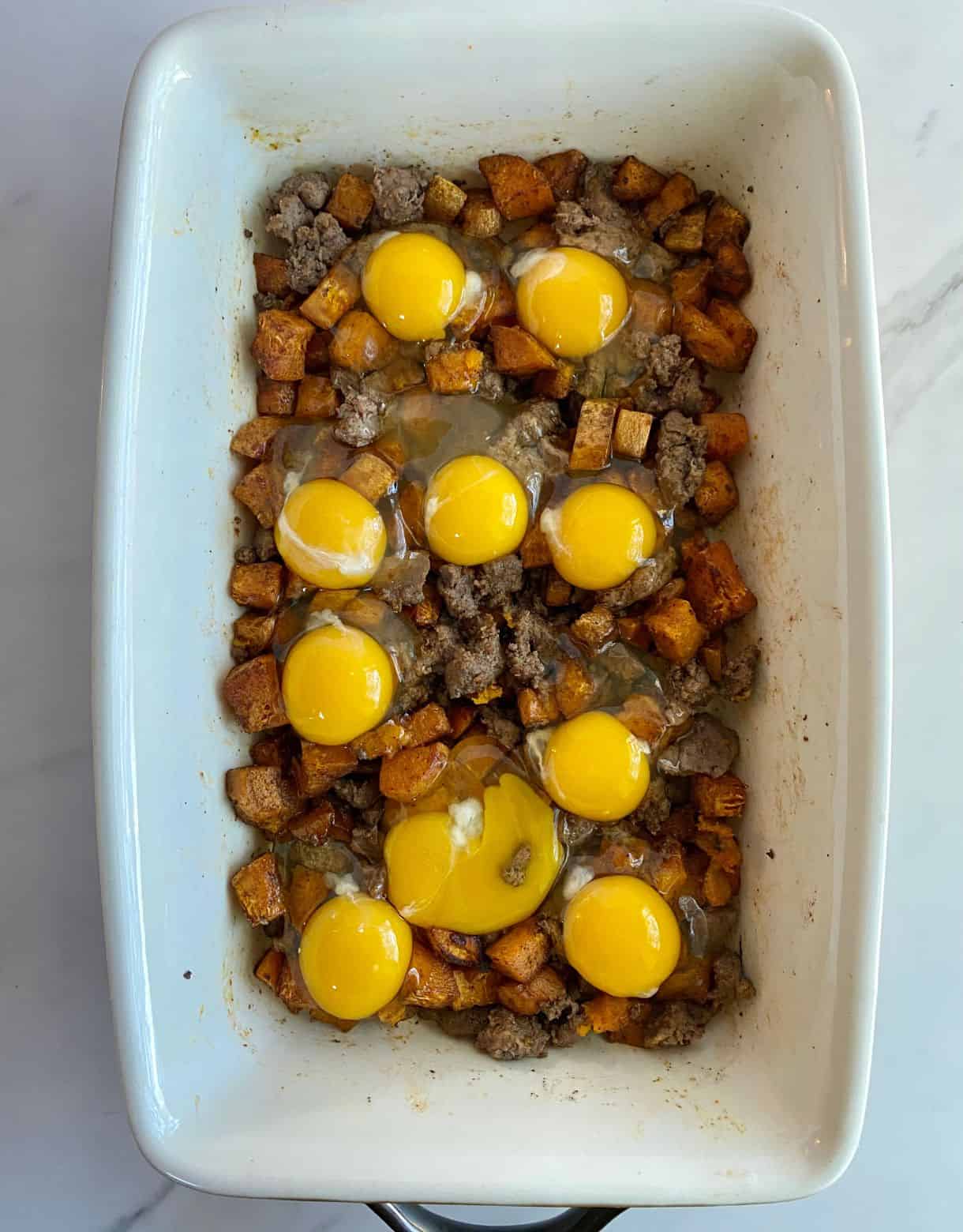 A baking dish of Sweet Potato Breakfast Hash with cooked sweet potatoes and sausage with raw eggs cracker over top.