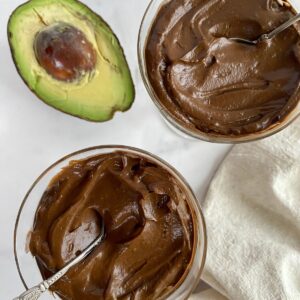 Two cups of Vegan Avocado Chocolate Mousse with spoons and half an avocado.