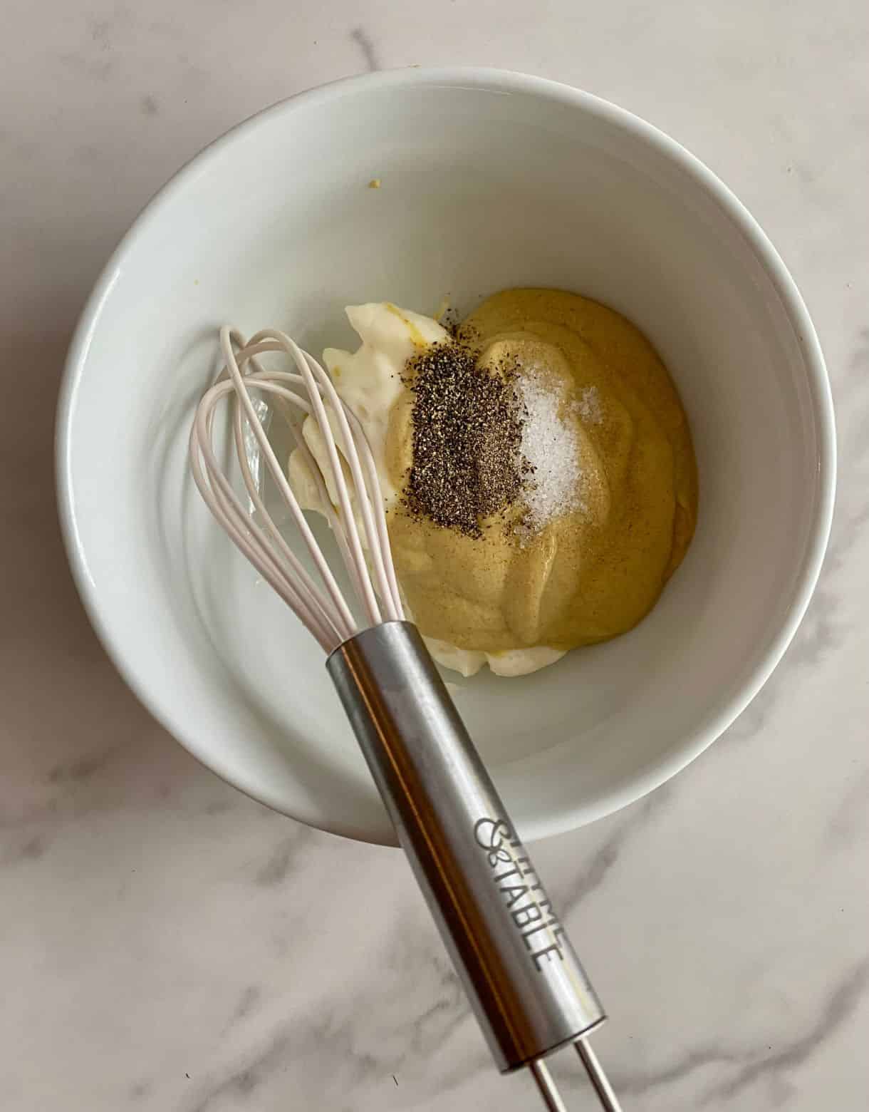 A bowl with mayonnaise, Dijon mustard, salt, pepper and a whisk.