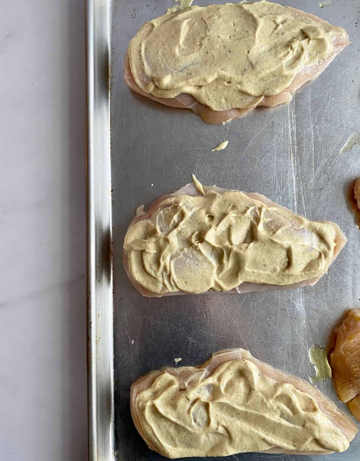 A sheet pan with raw chicken cutlets spread with a mixture of mayonnaise and Dijon mustard.