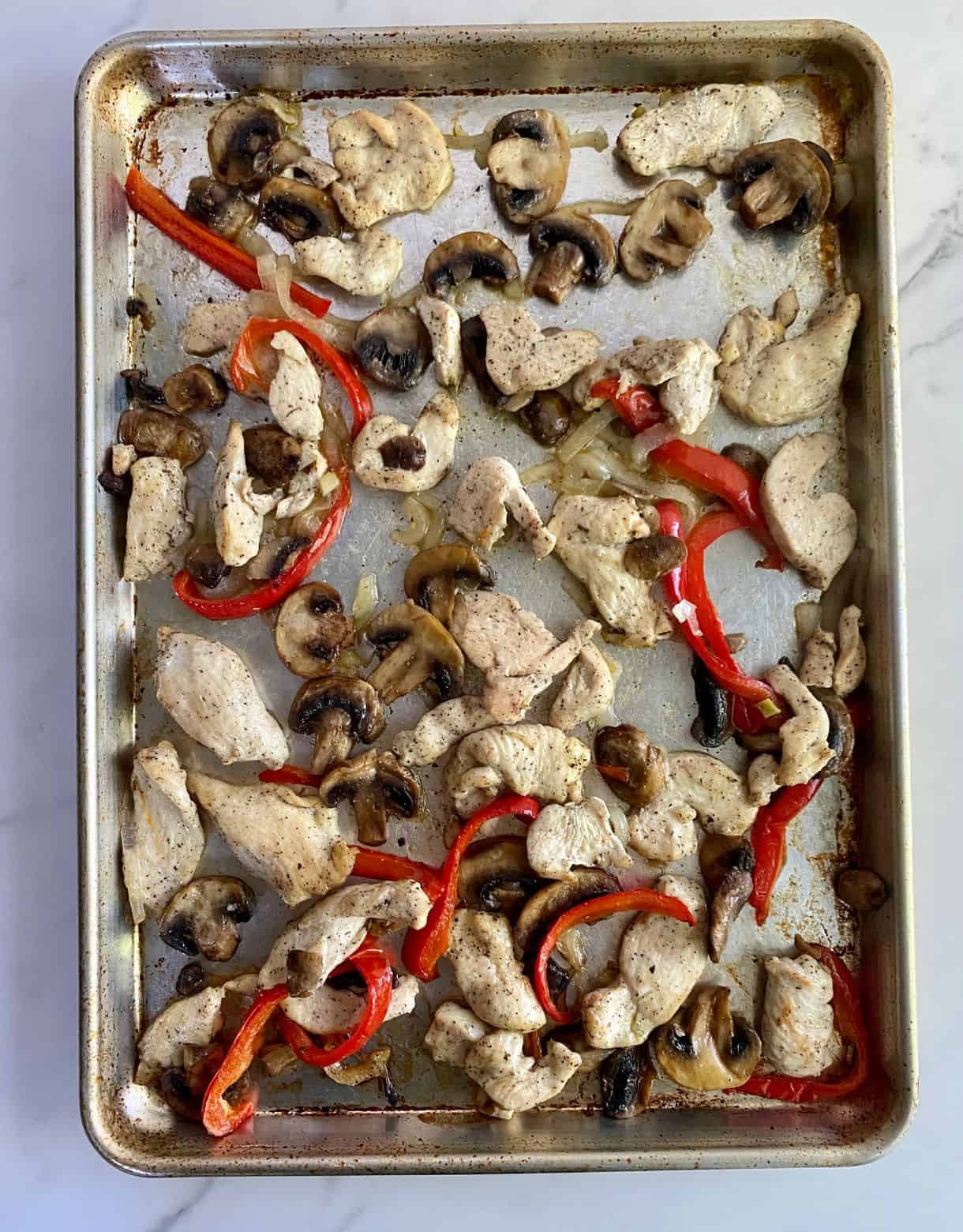 A baking sheet with cooked chicken, onion, mushroom and bell pepper.