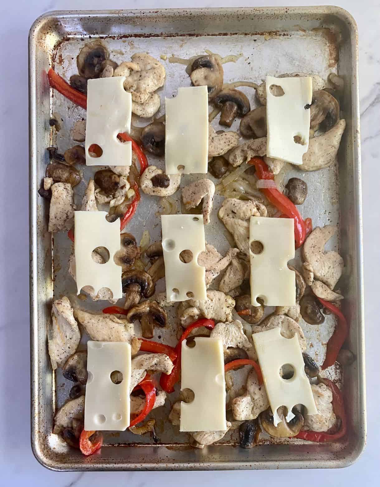 A sheet pan with cooked chicken, mushrooms, onions and bell pepper topped with slices of swiss cheese.