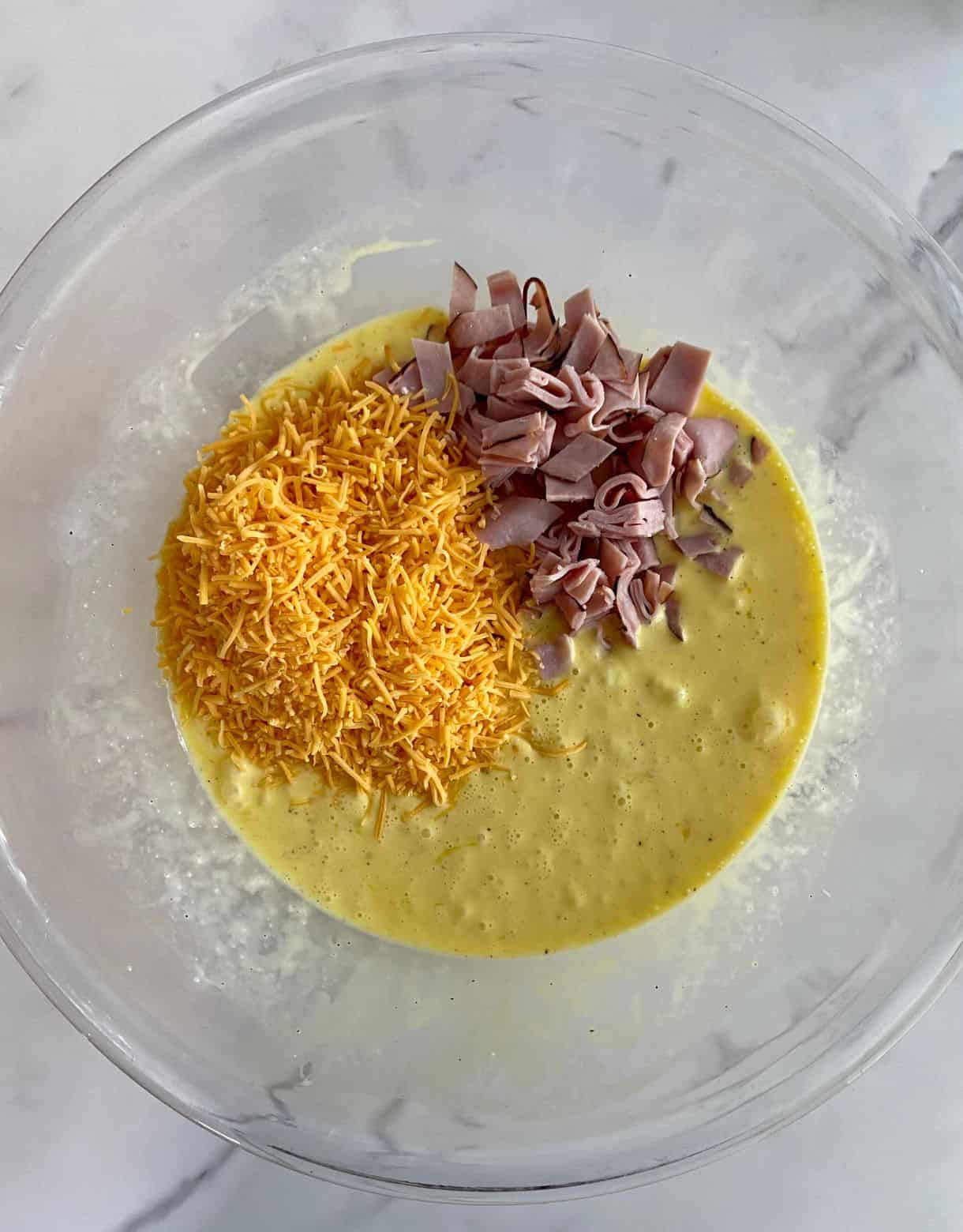 A bowl with whisked eggs and heavy cream with sliced ham and shredded cheddar cheese added but not yet stirred into the mixture.