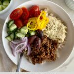 A bowl with cauliflower rice, seasoned grund beef, hummus, kalamata olives, siced red onion, chopped cucumbers, halved grape tomatoes and banana peppers.
