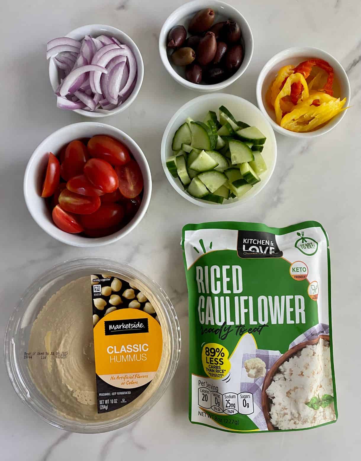 A bag of cauliflower rice, tub of hummus, halved grape tomatoes, sliced red onions, banana peppers, kalamata olives and diced cucumbers.