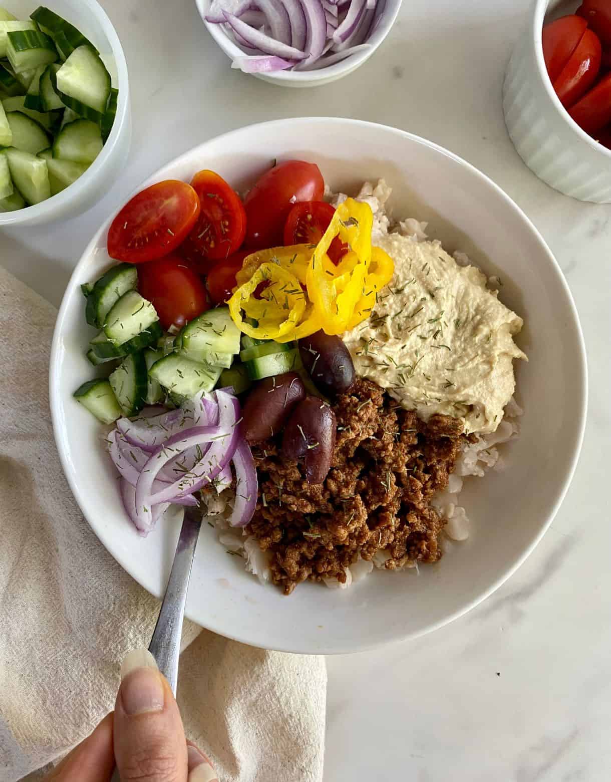 A bowl with cauliflower rice, seasoned grund beef, hummus, kalamata olives, siced red onion, chopped cucumbers, halved grape tomatoes and banana peppers.
