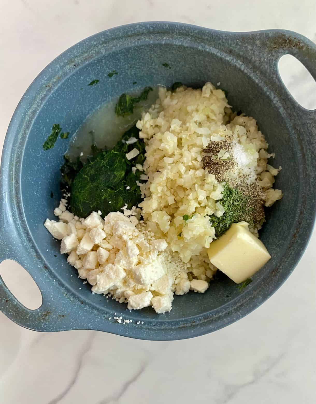 A dish with cauliflower rice, spinach, feta cheese, butter, salt, pepper, lemon juice and dill all added but not yet stirred.