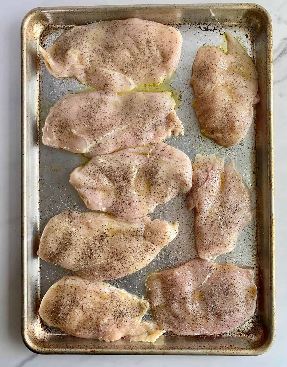 A sheet pan with raw chicken cutlets seasoned with salt, pepper and olive oil.