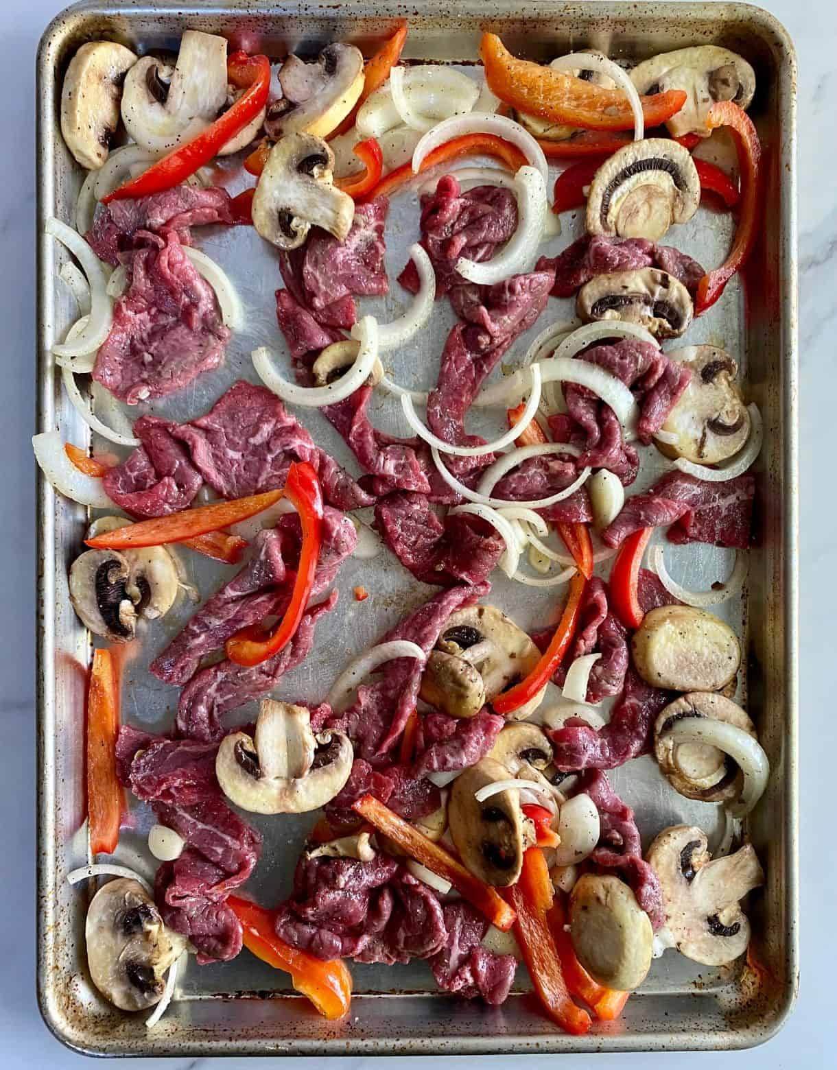 A sheet pan with sliced raw beef, mushrooms, onions and peppers ready to bake.