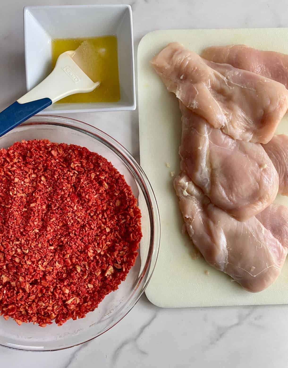 A cutting board with chicken cutlets, a dish of melted butter with a brush and a pan with crushed Takis.