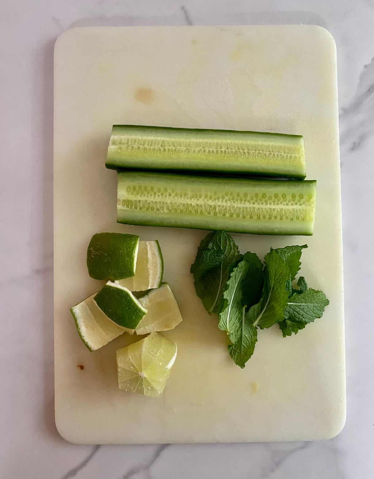 A cutting board with a cucumber sliced into quarters, a lime peels and some mint leaves.