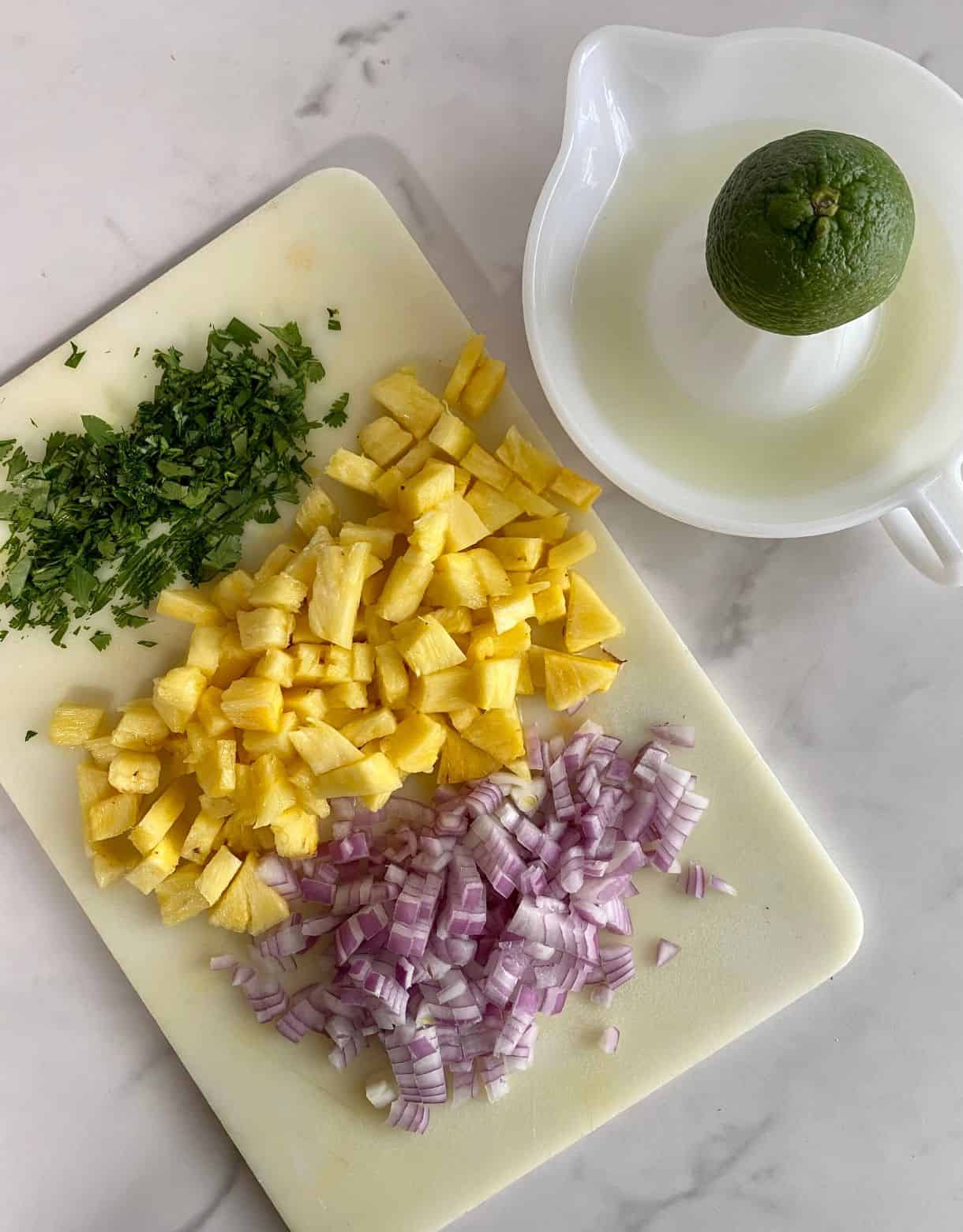 A juicer with half a lime on it and a cutting board with diced pineapple, diced red onion and chopped cilantro.