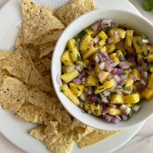 A bowl of Pineapple Pico de Gallo on a plate full of tortilla chips.