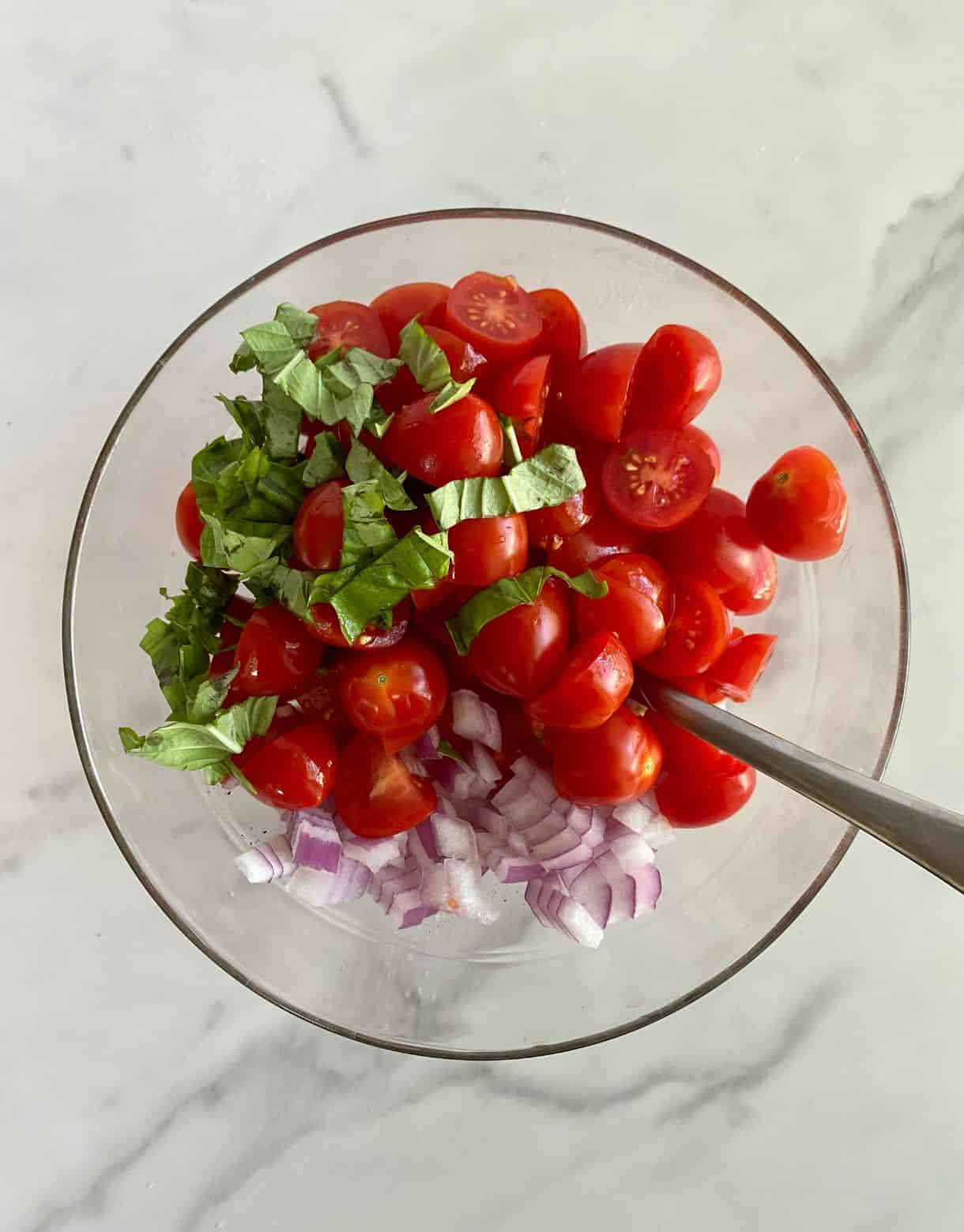 A bowl of halved grape tomatoes, chopped red onion, sliced basil, minced garlic, olive oil, salt and pepper ready to be stirred for Bruschetta.