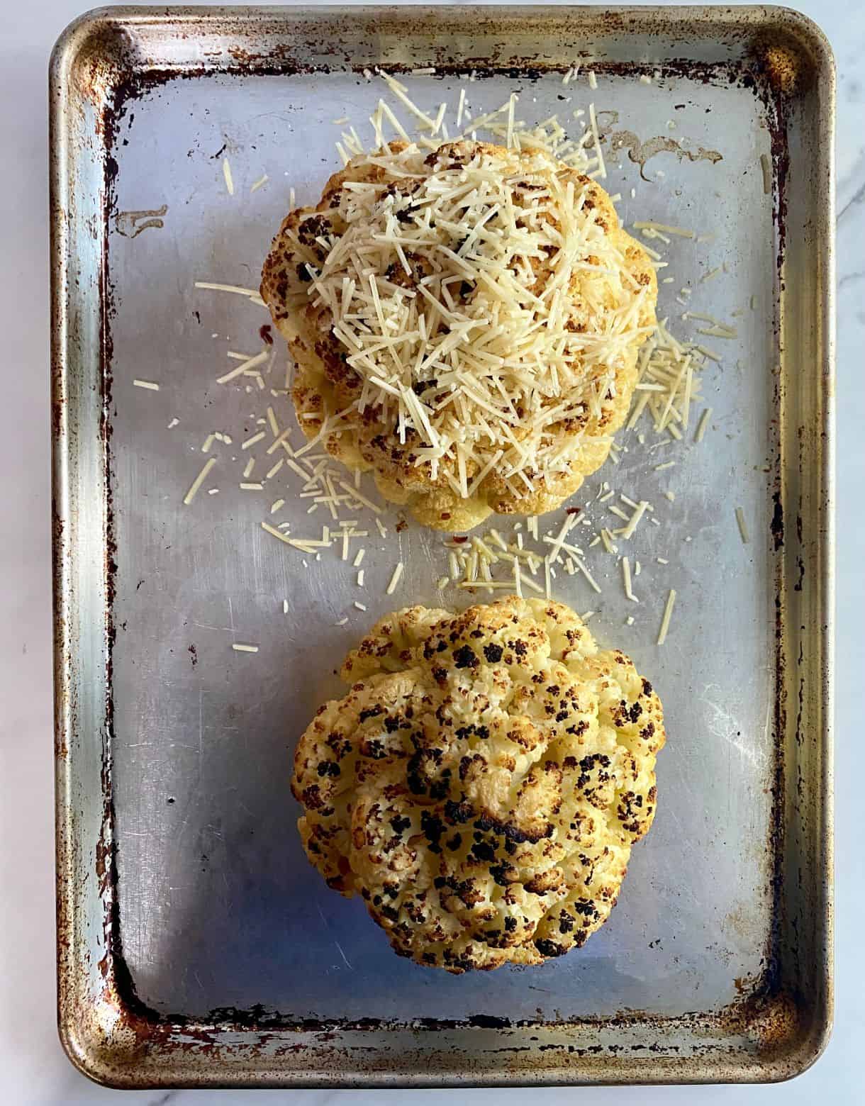 Two heads of whole roasted cauliflower nicely charred, one topped with shredded parmesan cheese.