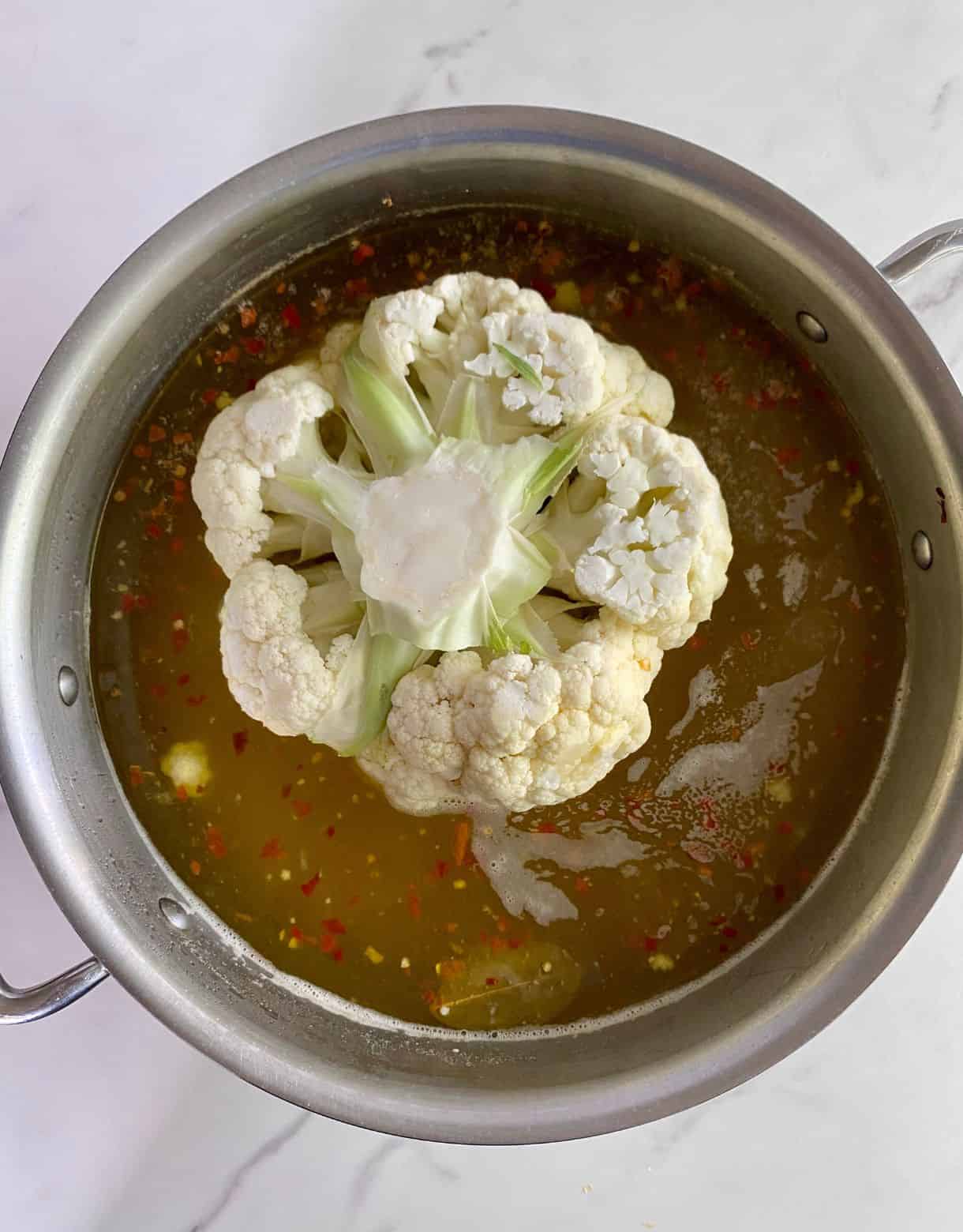 A pot of seasoned liquid with a whole head of raw cauliflower added to it.