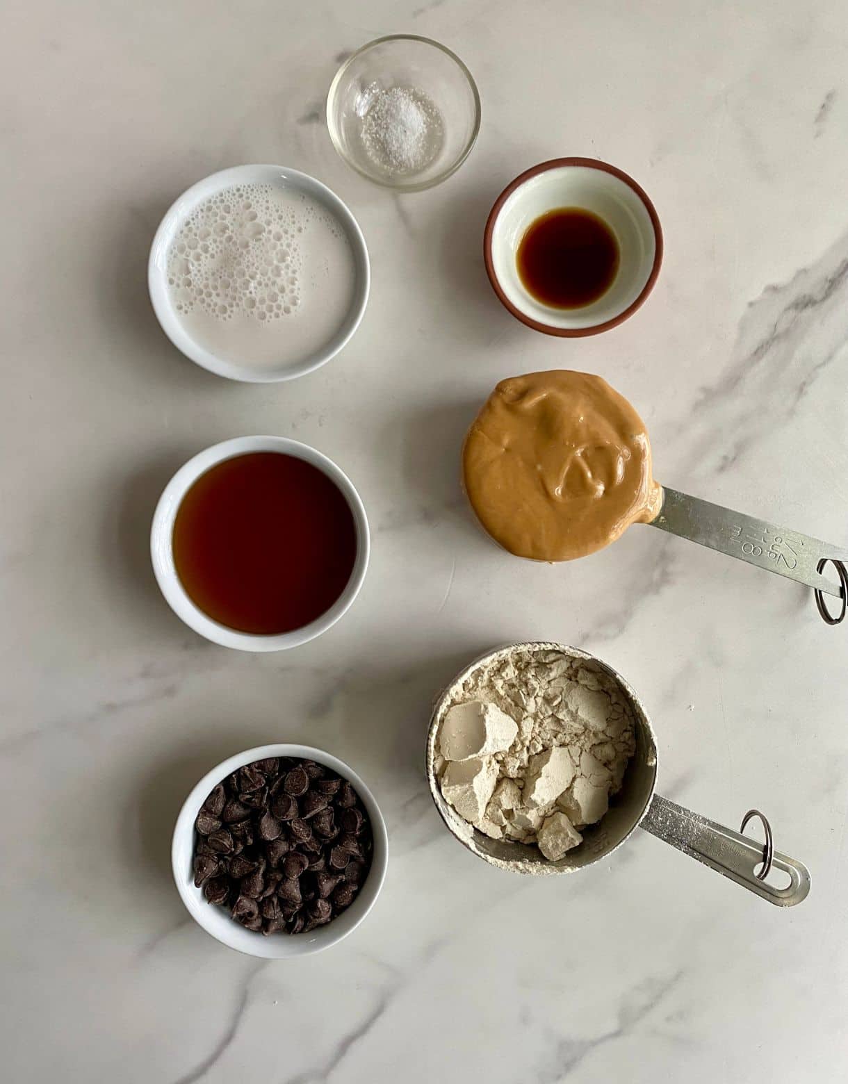 Small bowls of ingredients including protein powder, almond milk, peanut butter, chocolate chips, maple syrup, vanilla and salt.