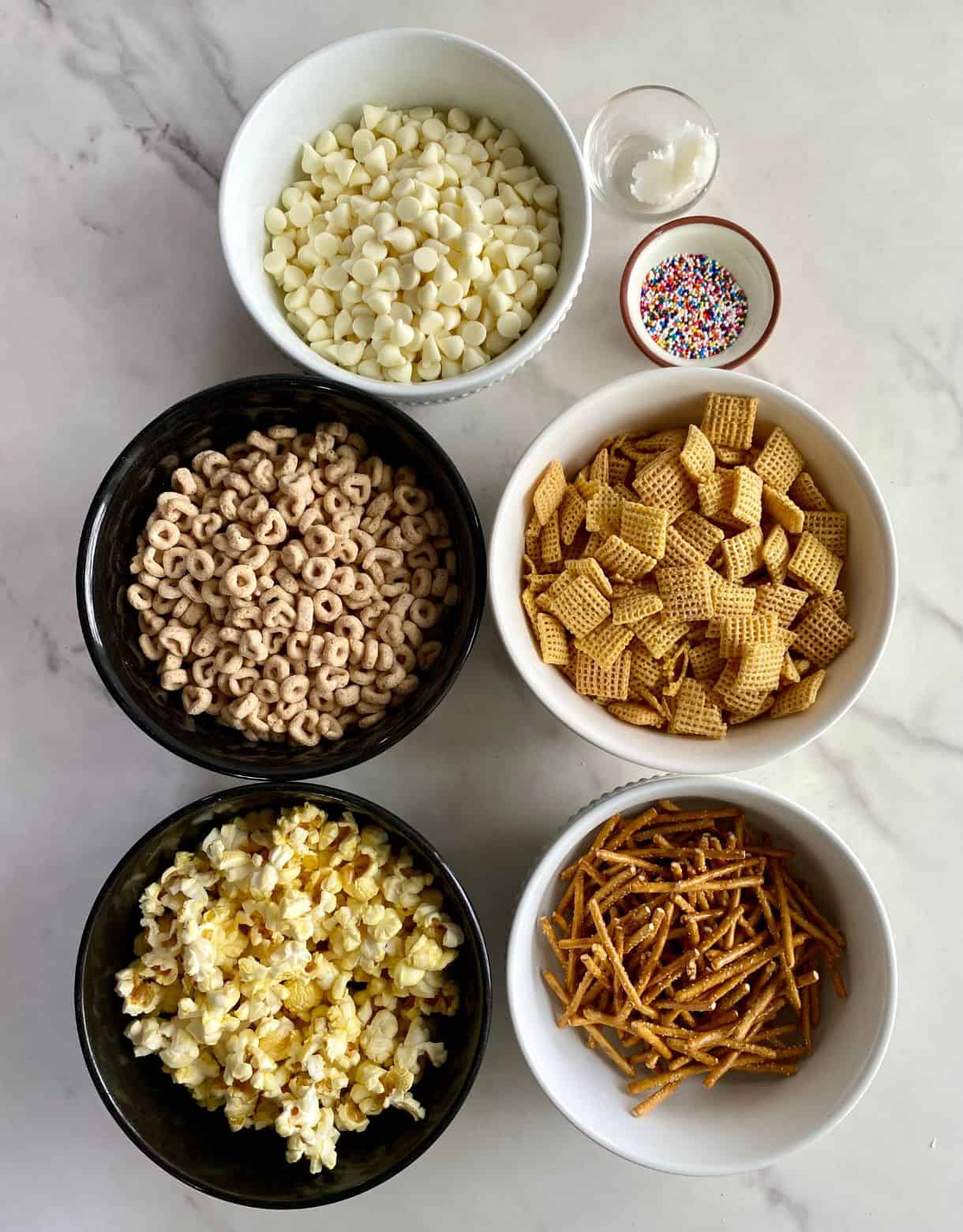 Small bowls of pretzels, white chocolate chips, coconut oil, sprinkles, cheerios, popcorn and corn chex.