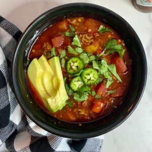 A bowl of cooked Slow Cooker Venison Chili topped with sliced avocado, chopped cilantro and sliced jalapenos.